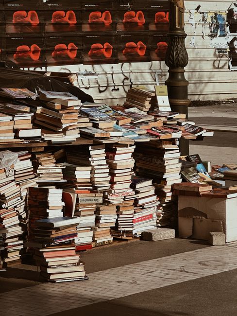 A large outdoor pile of books stacked in various heights on pavement, with some stacks leaning or uneven. The books are of different sizes, covers, and colors, including brown, black, white, and red,